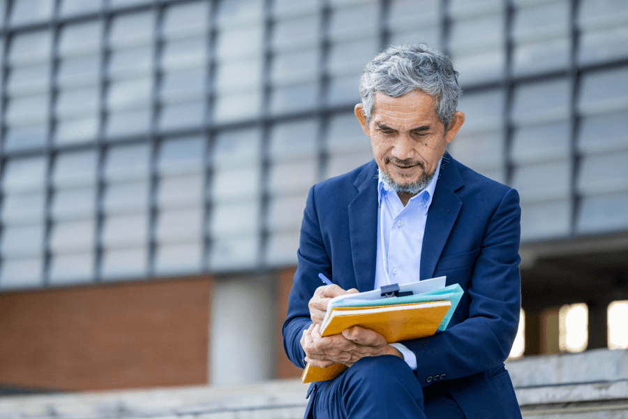 Older man in a navy suit sits outdoors on steps, writing on documents clipped to a stack of colorful folders. He appears focused, with a modern office or institutional building in the background.