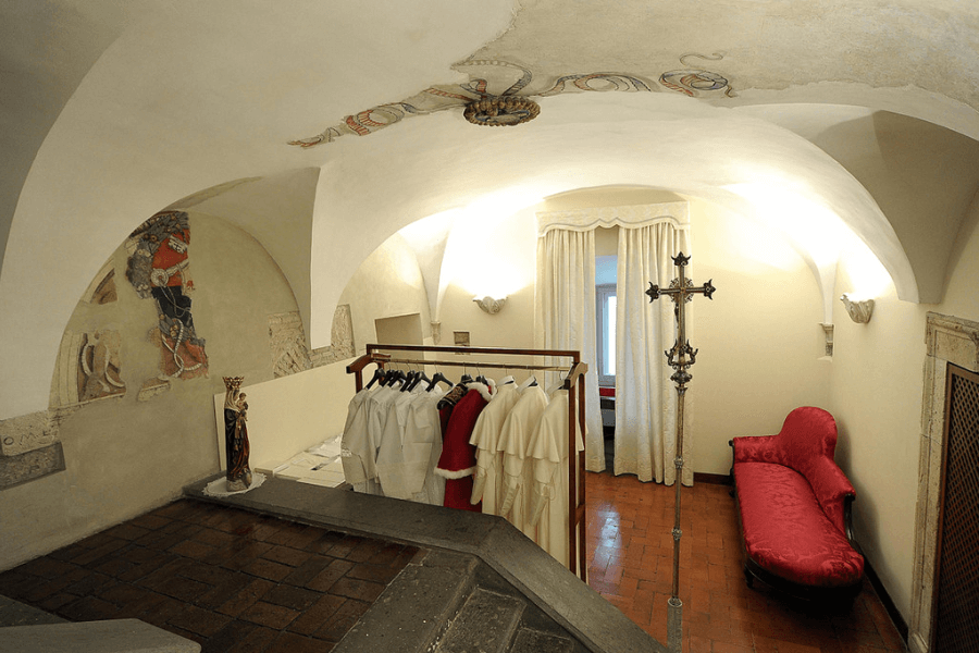 A well-lit, arched dressing chamber with religious garments on a clothing rack, including white and red robes. The room features a red velvet chaise lounge, a tall ornate cross, wall murals, and a statue of the Virgin Mary, suggesting a ceremonial or papal setting.