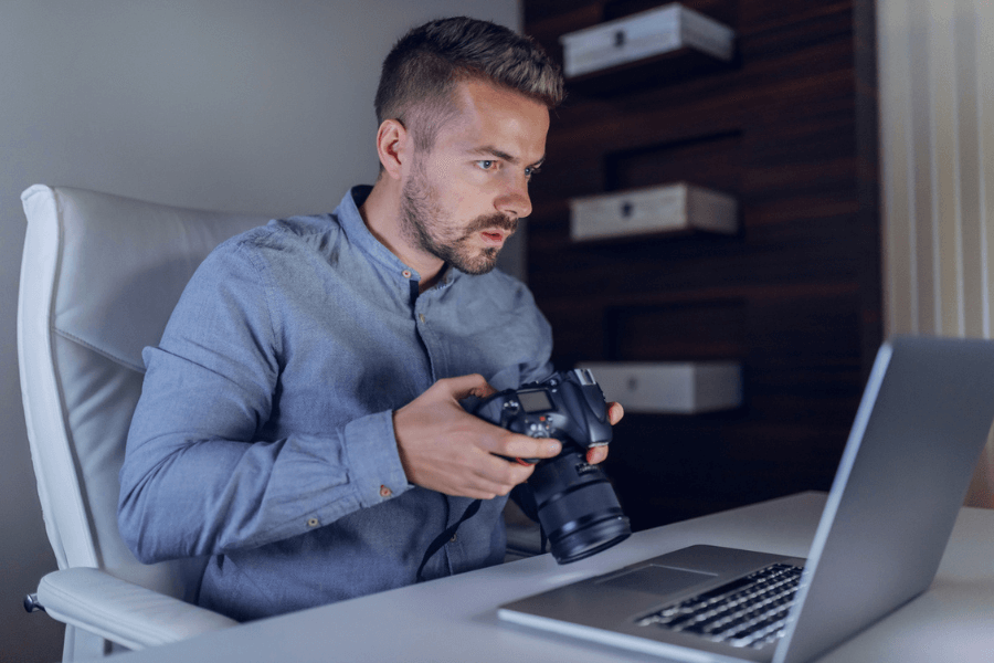 Man in a button-up shirt sits at a desk holding a DSLR camera, looking intently at his laptop screen. The scene suggests he's reviewing or editing photos in a focused home or studio workspace.