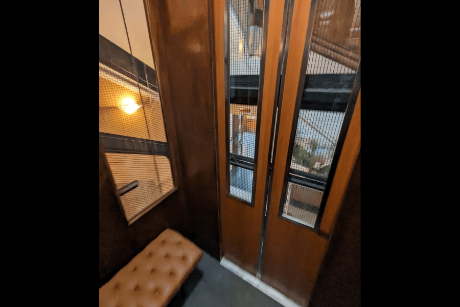 Interior of a vintage elevator with wooden paneling, a tufted leather bench, and narrow windows in the door with mesh screens showing the stairwell outside. A mirror on the wall reflects the dim lighting and part of the stair railing.