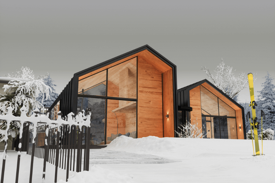 Modern wooden cabin with large glass windows set in a snowy landscape, surrounded by snow-covered trees and a black metal fence. A pair of bright yellow skis and poles stand upright in the snow near the entrance, suggesting a cozy ski retreat.