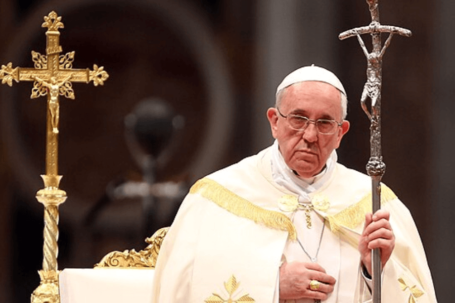 Pope Francis stands solemnly during a liturgical ceremony, wearing white and gold vestments and holding a silver papal ferula topped with a crucifix. A golden processional cross is visible beside him, set against a blurred church interior.