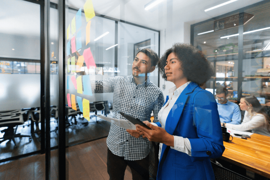 Two professionals collaborate in a modern office, standing in front of a glass wall covered with colorful sticky notes. The man gestures while discussing ideas, and the woman in a blue blazer holds a tablet, actively engaged in planning.