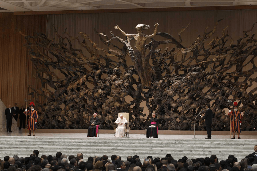 Pope presides over a public audience in the Vatican’s Paul VI Audience Hall, seated before the massive bronze “Resurrection” sculpture depicting Christ rising from chaos. Swiss Guards in colorful uniforms stand at both sides of the stage, while clergy and guests surround the Pope.