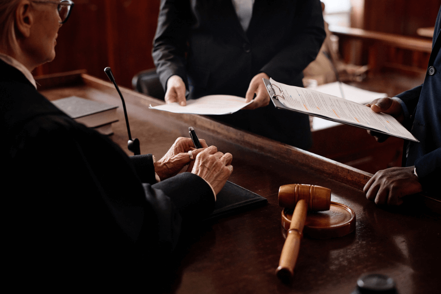 Close-up of a judge signing a document in a courtroom, with two attorneys holding clipboards and presenting paperwork. A gavel rests on the wooden bench, emphasizing the formal and legal setting.
