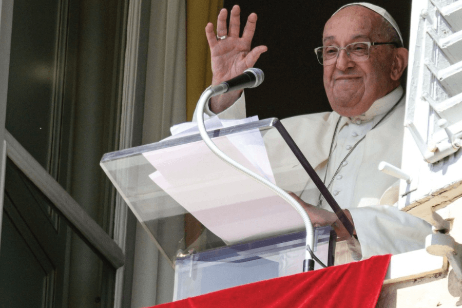 Pope Francis smiles and waves from a window balcony, standing behind a clear lectern with papers and a microphone during a public address. A red cloth hangs from the ledge, signifying an official Vatican appearance.