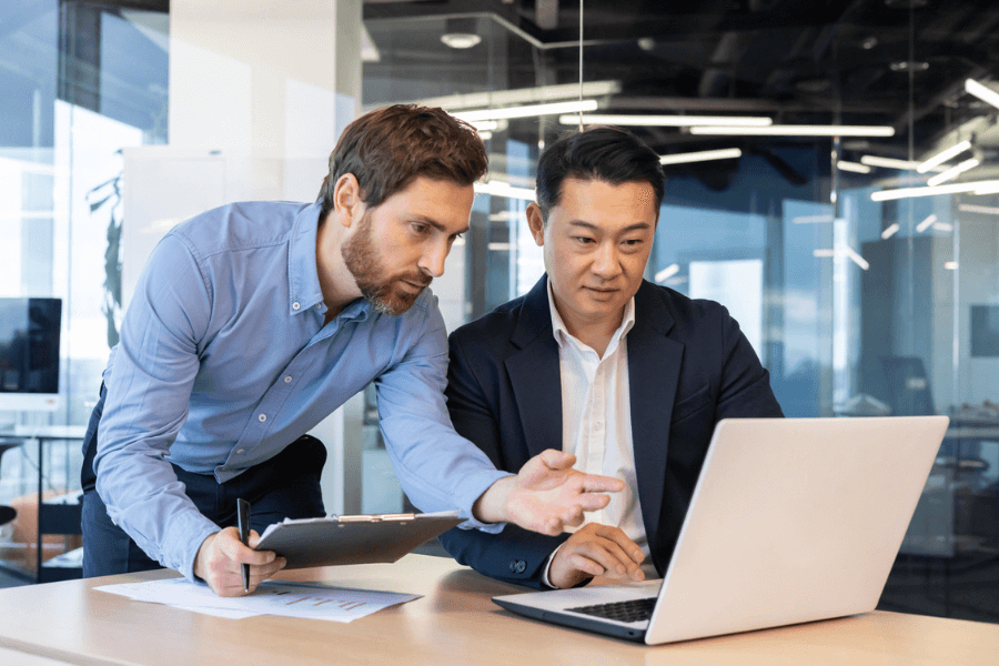 Two businessmen collaborate at a desk in a modern office; one points at a laptop screen while holding a clipboard, and the other listens attentively. The workspace is well-lit with glass walls and contemporary lighting fixtures.