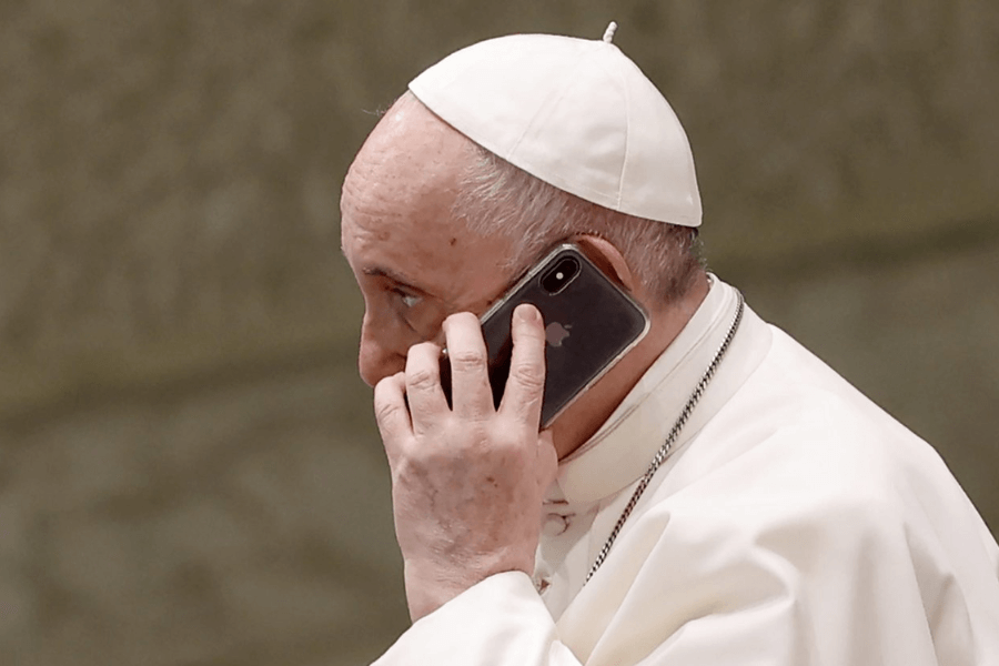 Pope Francis, dressed in his white papal robes and skullcap, holds a smartphone to his ear while engaged in a phone conversation. The image captures a candid and modern moment of communication.