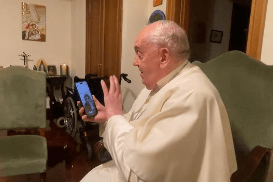 Pope Francis sits in a cozy, dimly lit room, smiling and waving during a video call on a smartphone. A wheelchair is visible in the background, along with religious decorations and lit candles on a side table.