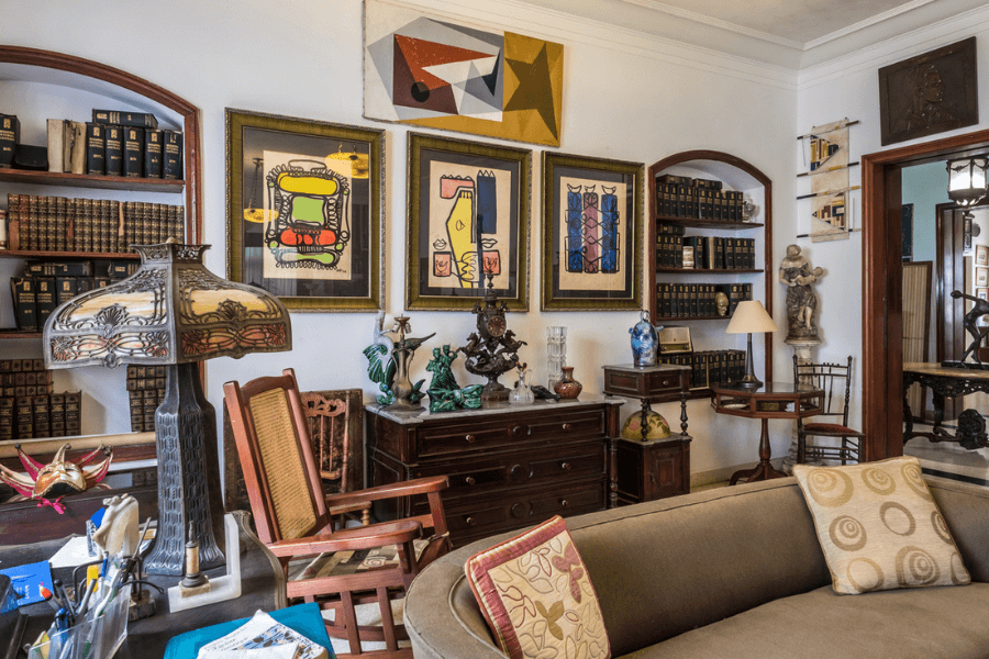 Eclectic and cozy living room filled with antique furniture, colorful abstract artwork, and decorative collectibles. Shelves lined with old books and unique sculptures give the space a vintage, intellectual charm.