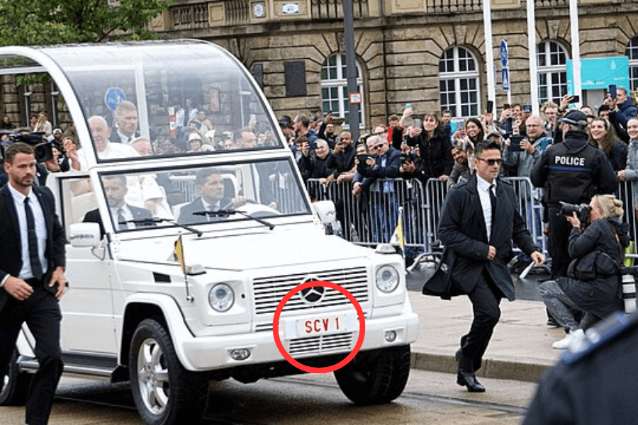 Pope Francis waves from within a white Popemobile with a transparent protective canopy during a public procession. The license plate reads “SCV 1,” denoting “Status Civitatis Vaticanae,” the official registration for the Vatican City’s lead vehicle. The crowd behind barricades eagerly takes photos while security personnel surround the vehicle.
