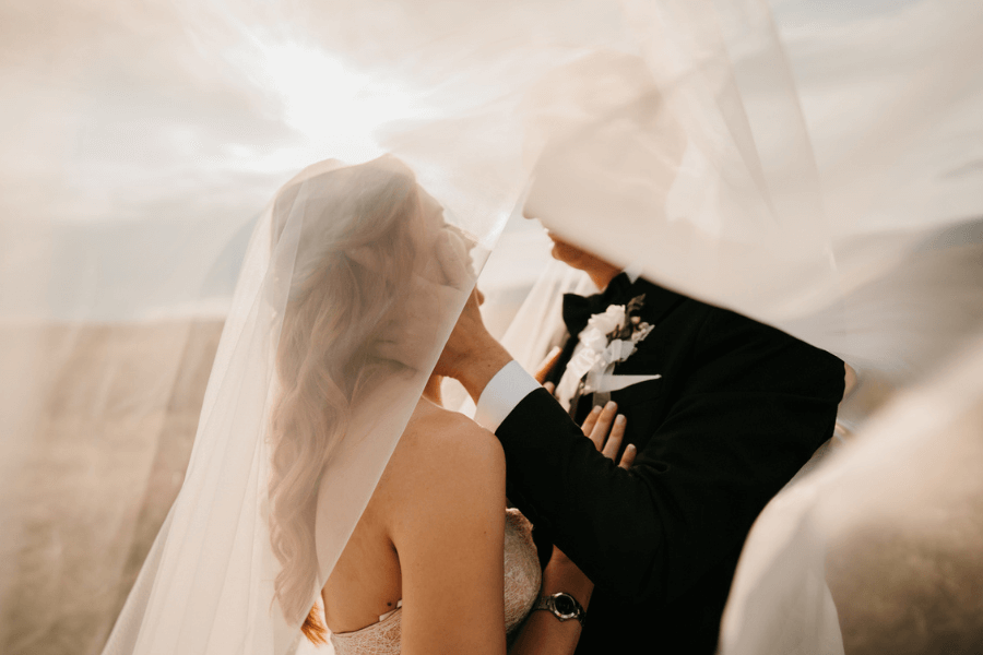 Bride and groom share an intimate moment under a flowing veil during golden hour, with soft sunlight illuminating the scene. The groom gently touches the bride’s face as they stand close, dressed in formal wedding attire.