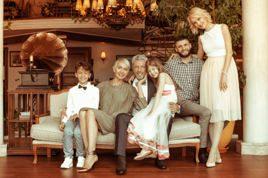Stylishly dressed family poses together in an elegant, vintage-style living room with warm lighting, a grand chandelier, and a gramophone. The group includes smiling adults and children, exuding a sense of togetherness and celebration.