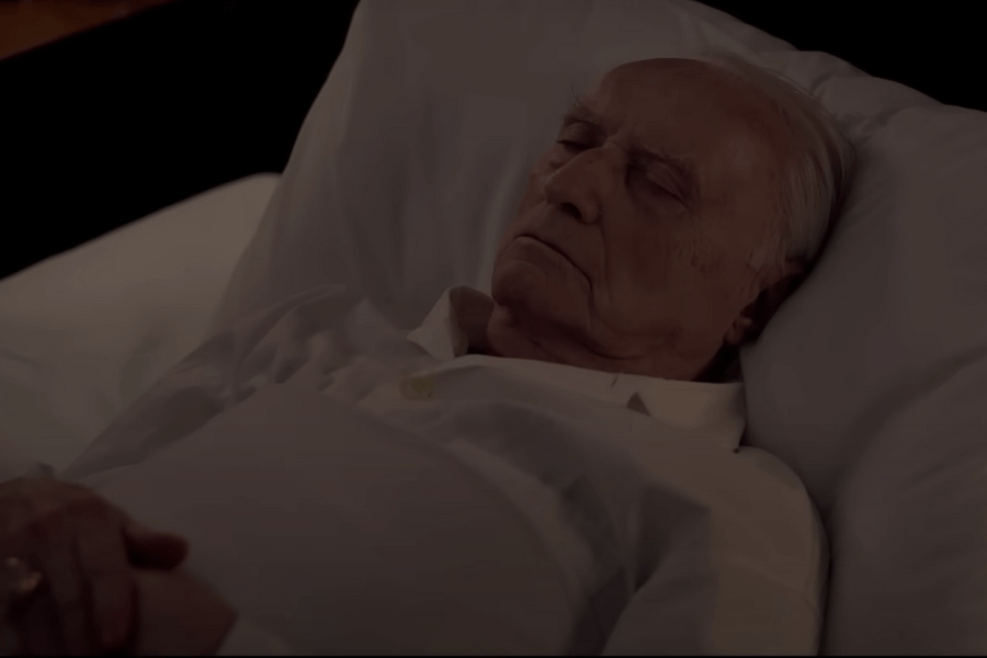An elderly man lies peacefully with hands clasped, eyes closed, and dressed in a white shirt, resting on a pillow in a dimly lit room. The solemn expression and setting suggest the depiction of a final moment or a scene of repose.