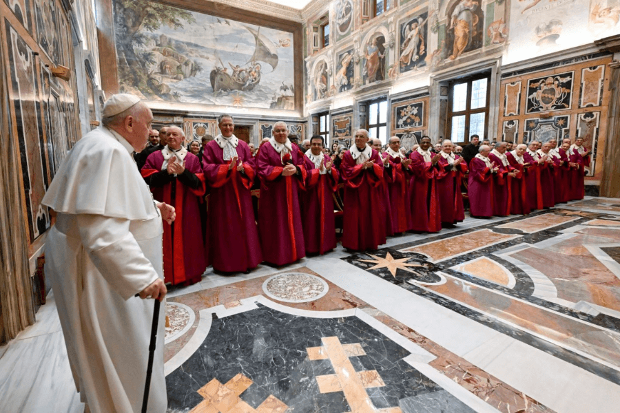 Pope Francis, walking with a cane, greets a line of clergy dressed in maroon and white formal ecclesiastical robes inside a richly decorated Vatican hall with marble floors and frescoed walls. The group stands in formation, applauding the Pope as he enters.