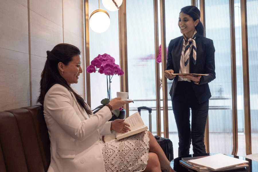 Smiling businesswoman in a white suit receives a cup of coffee from a hotel or lounge attendant in a dark suit with a neck scarf, while sitting with a book and luggage beside her. Elegant orchids and a refined setting suggest a premium hospitality environment.