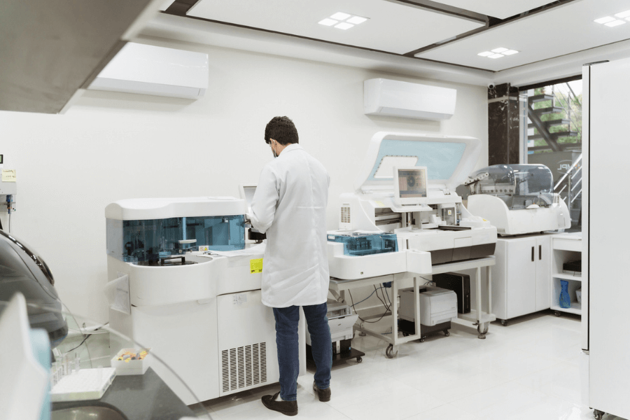 A lab technician wearing a white coat operates advanced diagnostic equipment in a modern laboratory setting, surrounded by machines and clinical testing devices. The environment is clean, bright, and organized, with white walls and glossy floors.