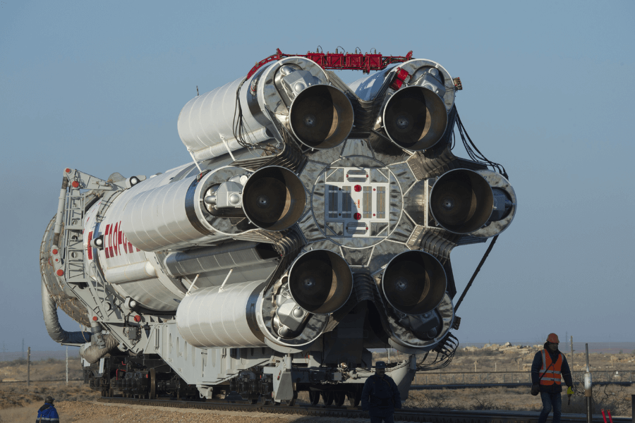 A large space rocket, viewed from the rear to highlight its powerful engines, is being transported horizontally on a rail-mounted platform across a barren landscape. Several workers in safety gear walk alongside the rocket during what appears to be a pre-launch transport or positioning operation.