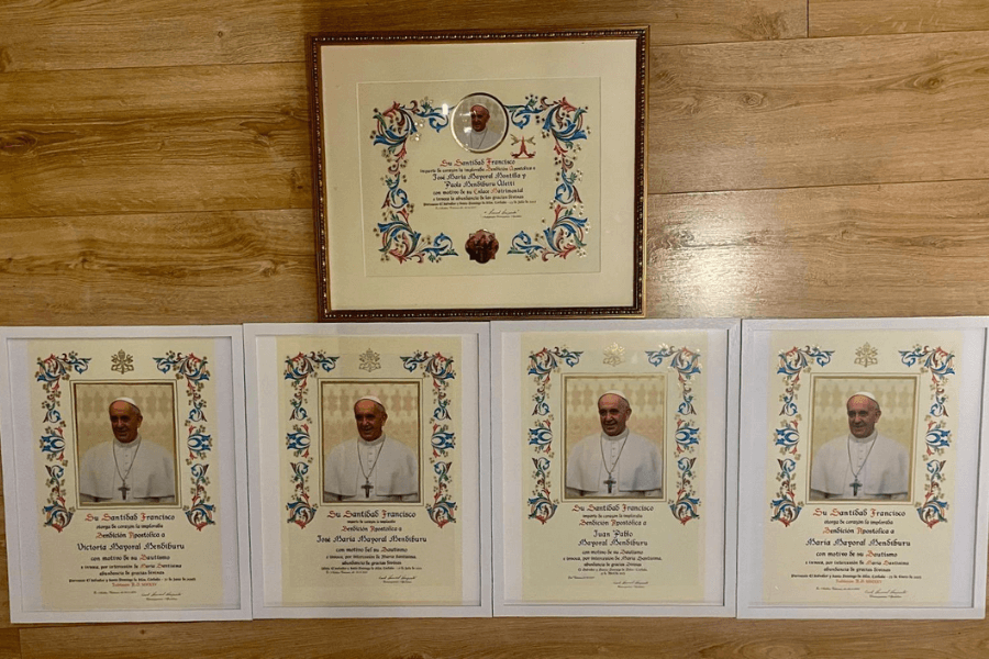 Five ornate Papal Blessing certificates from Pope Francis are displayed on a wooden floor, each bordered with colorful floral patterns and bearing a photo of the Pope. Four certificates are unframed and laid side by side, while one is framed and centered above them.