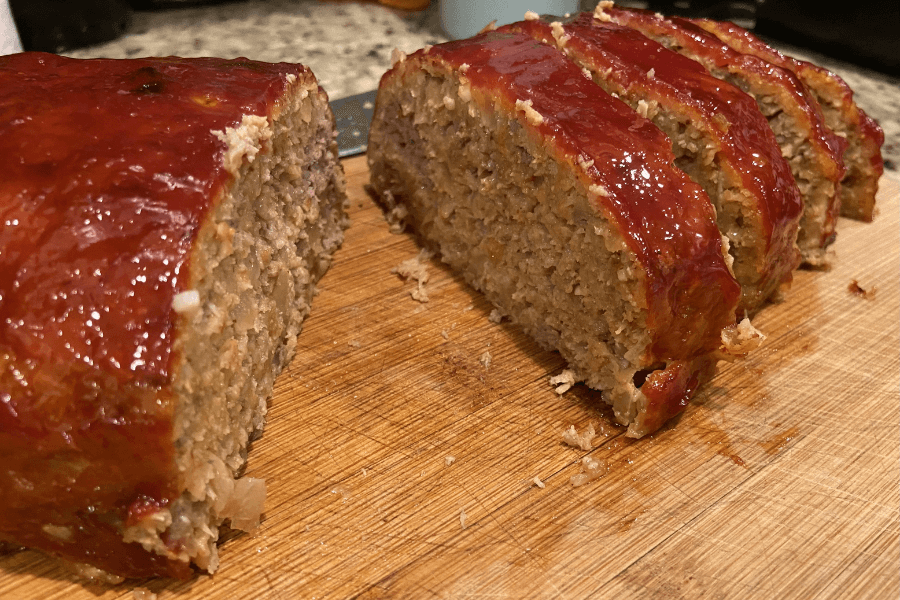 A freshly sliced meatloaf on a wooden cutting board, topped with a glossy ketchup glaze. The interior reveals a moist, well-mixed blend of ground meat, onions, and breadcrumbs.