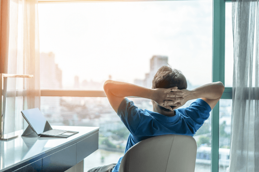Man in a blue shirt sits at a modern desk with a laptop, leaning back with hands behind his head while gazing out a large window at a bright city skyline. The setting suggests a relaxed work-from-home or remote work environment.