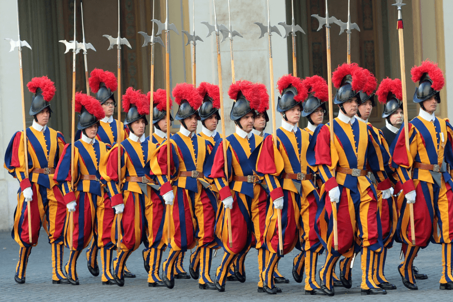 A formation of Swiss Guards marches in traditional Renaissance-style uniforms of blue, red, and yellow stripes, with red feathered plumes on their black helmets. Each guard holds a tall halberd while walking in sync during a ceremonial display.