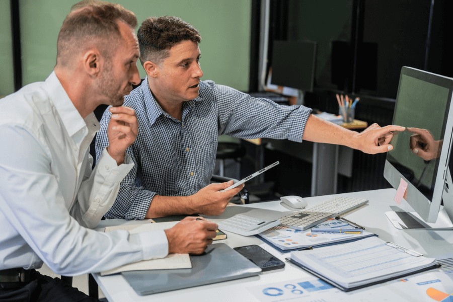 Two men in business attire working together at a desk, with one pointing at a desktop monitor while the other takes notes. The workspace is filled with charts, reports, a tablet, and office supplies, suggesting a collaborative planning or analysis session.