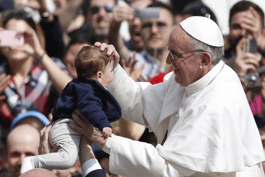 A smiling Pope in white papal robes and skullcap gently places his hand on a baby's head while someone lifts the baby toward him. The joyful crowd in the background captures the moment with their phones, highlighting the celebratory atmosphere.