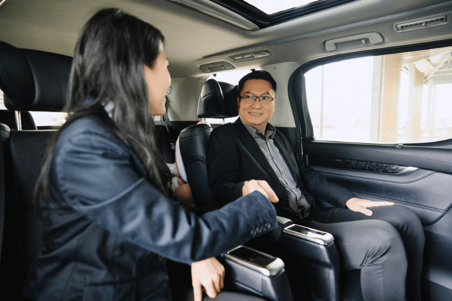 Two business professionals in formal attire shake hands while seated in the back of a luxury vehicle, smiling and engaged in conversation. The setting suggests a successful meeting or partnership, with natural light coming through the car windows.