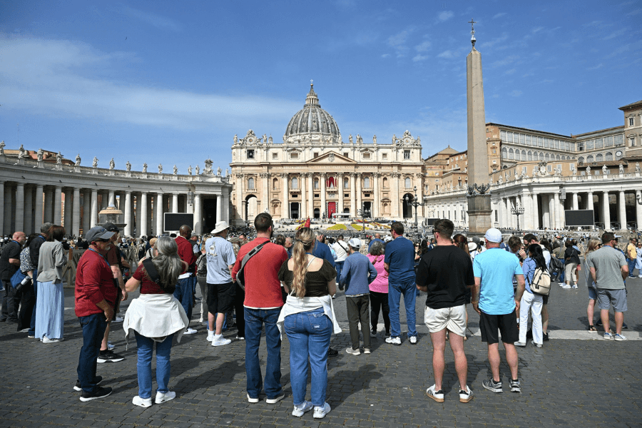 Crowds of tourists and pilgrims gather in St. Peter's Square in Vatican City, facing the grand facade of St. Peter's Basilica under a bright blue sky. The central obelisk and the colonnades surrounding the square add to the iconic setting.