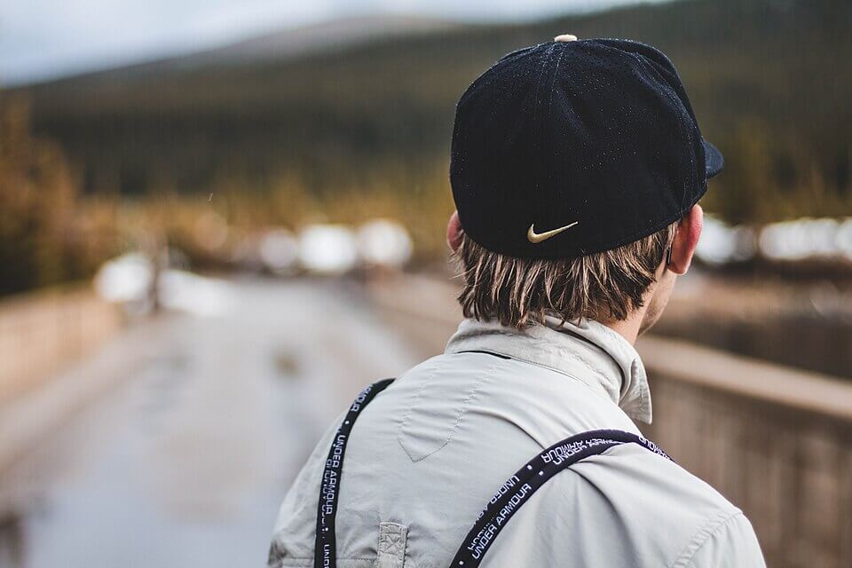 A person with light brown, slightly wet hair is seen from behind, wearing a black Nike cap and a light gray collared shirt. They have dark Under Armour branded suspenders, and they are standing on a wet path surrounded by blurred trees and mountains in the background.