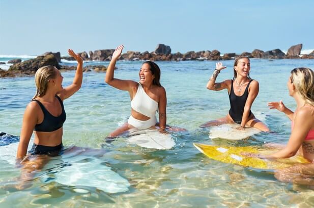 Group of young women in swimwear sit on surfboards in clear ocean water. Friends enjoy sunny beach, have fun, high-five. Girls at tropical surf camp, bond, laugh, ocean adventure.
