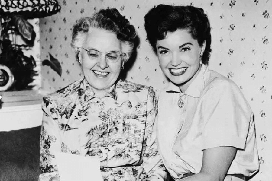 Black and white photo of two smiling women seated side by side. The older woman on the left wears glasses and a patterned dress, looking down at a piece of paper, while the younger woman on the right, with dark, styled hair and a bright smile, wears a short-sleeved blouse with a brooch at the collar. They appear to be enjoying a light-hearted moment in a mid-century home setting.