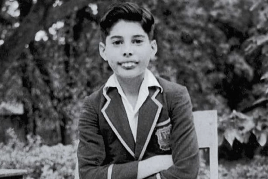 Black and white photo of a young boy sitting on a wooden chair outdoors, dressed in a formal school blazer with white piping and an embroidered crest. He has neatly combed hair and is looking directly at the camera with a composed expression, arms casually crossed.