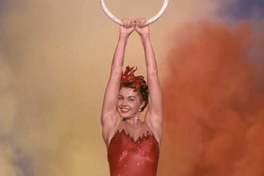 Color image of a smiling female performer in a sparkly red costume and headpiece, hanging from a suspended ring with both arms raised. The background is filled with vibrant orange and yellow stage smoke, suggesting a circus or theatrical performance.