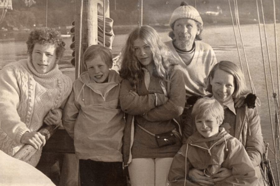 Sepia-toned photo of a smiling family of six aboard a sailboat, dressed in cozy sweaters and jackets. The group includes two adults and four children, posing close together with a calm body of water and wooded shoreline in the background.