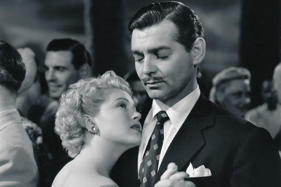 Black and white film still of a glamorous couple dancing closely. The woman with short, curled blonde hair gazes up longingly at the man, who has slicked-back dark hair and a thin mustache, wearing a suit and patterned tie. They’re surrounded by a softly focused crowd at a formal gathering.