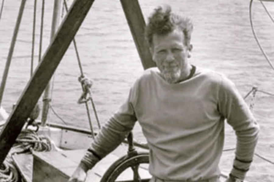 Black-and-white photo of a man with tousled hair and a short beard, standing at the helm of a sailboat. He’s wearing a crewneck sweater and leaning casually on the ship’s wheel, with ropes and rigging visible behind him and calm water in the background.