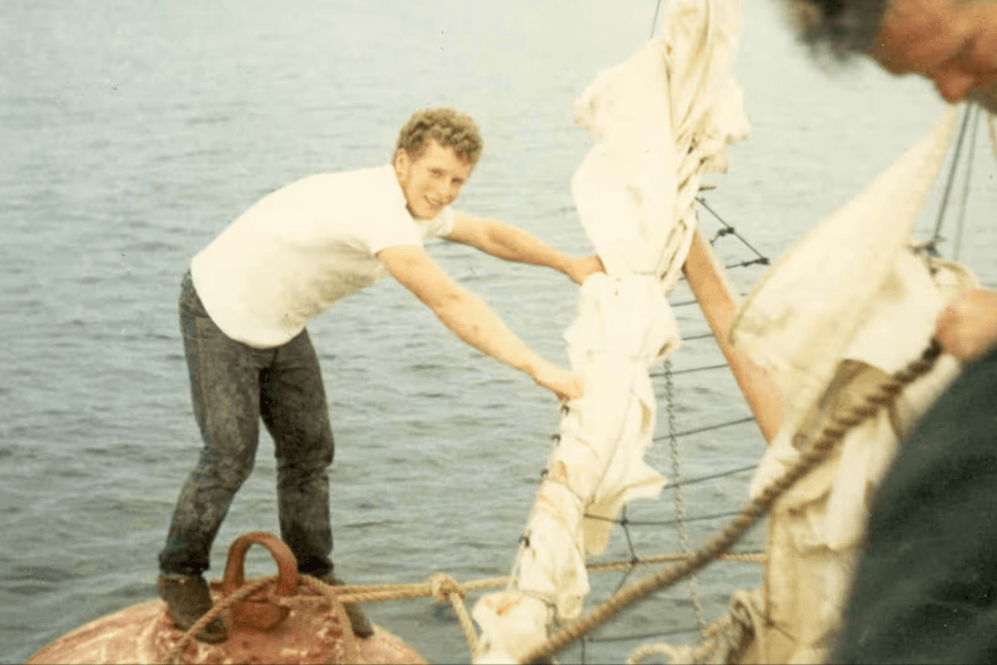 Vintage color photo of a young man in a white T-shirt and dark pants standing on a large red buoy at sea. He smiles toward the camera while holding a sail that is being passed from a nearby boat, with another person partially visible on the right helping manage the rope.