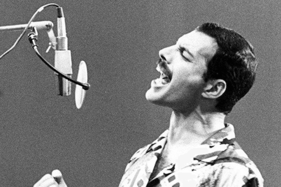 Black and white photo of Freddie Mercury passionately singing into a studio microphone, wearing a patterned shirt. His head is tilted back slightly, eyes closed, and fist clenched, capturing the intensity and emotion of his vocal performance.
