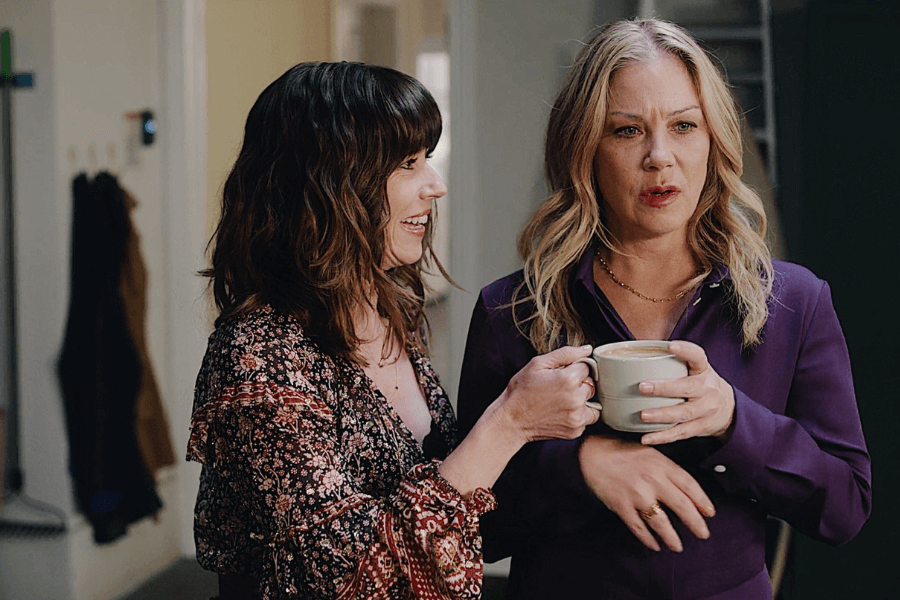 Two women stand indoors sharing a moment—one wearing a floral dress smiles warmly while offering a cup of coffee to the other, who looks surprised or mid-sentence in a purple blouse. The setting appears to be a cozy home or apartment.