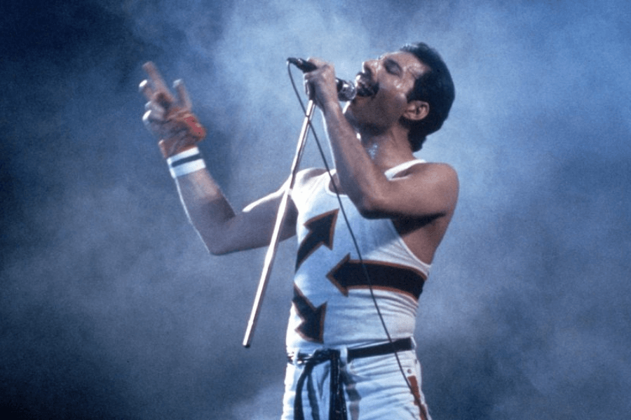 Freddie Mercury performing live on stage in a white tank top with red and black arrow designs, passionately singing into a microphone while surrounded by dramatic stage smoke. He wears sweatbands on his wrist and holds the mic stand with intensity, captured mid-performance.