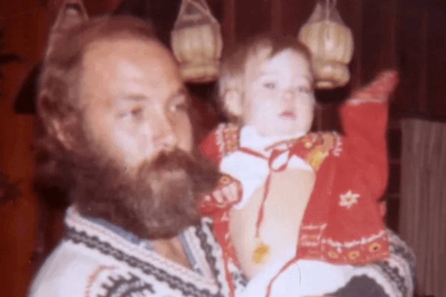 A vintage photo of a bearded man wearing a patterned sweater holding a baby dressed in a red outfit with embroidered details. The background includes wooden walls and hanging wicker lamps, suggesting a cozy, rustic interior.
