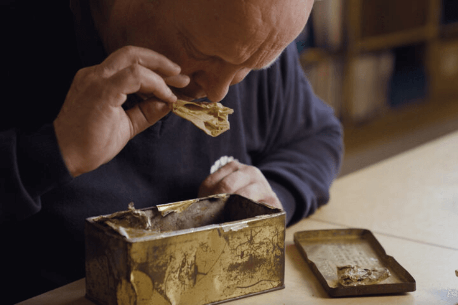Close-up of a bald man closely sniffing a preserved piece of food held in his hand, taken from a dented, partially opened golden tin can on the table. Another piece of the same food rests on the removed lid beside the can, suggesting examination of long-stored rations.