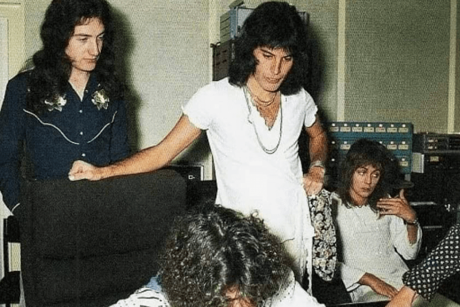 Candid photo of Queen band members inside a recording studio, with Freddie Mercury in a white shirt standing at the center, leaning on a chair. John Deacon and Roger Taylor are nearby, with Brian May sitting and focusing on something out of view, surrounded by vintage audio equipment and recording consoles.
