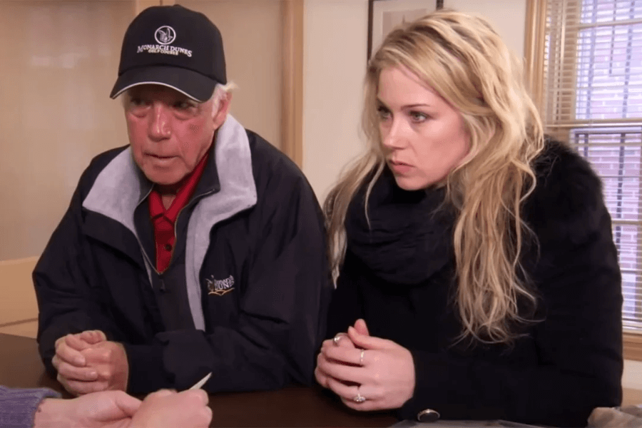 A serious-looking woman with long blonde hair sits beside an older man wearing a black cap and jacket. They are at a table in what appears to be an office or consultation room, listening intently to someone out of frame.