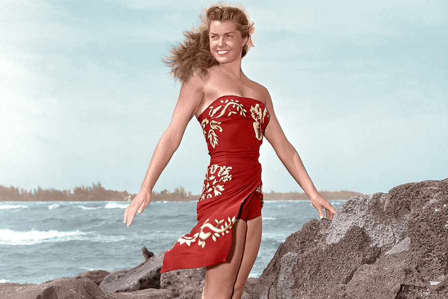 Color photo of a smiling woman standing on volcanic rocks by the ocean, wearing a strapless red sarong-style outfit with yellow floral designs. Her hair is windblown, and the waves crash behind her under a lightly overcast sky, evoking a tropical island vibe.
