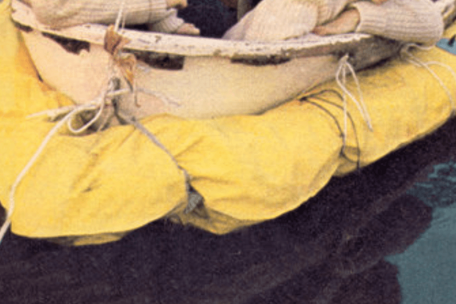 Close-up image of the lower portion of a small, weathered lifeboat resting atop a bright yellow inflatable flotation collar. The boat appears secured with multiple ropes, and a person's folded arms in a sweater are visible just above the rim.