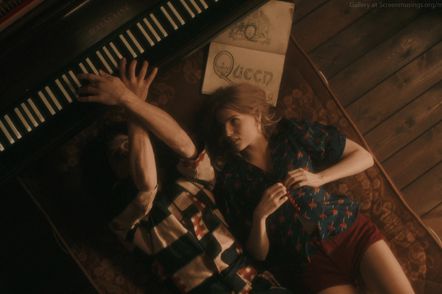 Cinematic scene showing a man and woman lying on a vintage patterned couch beside a piano, with the man reaching up toward the keys. A sketchbook labeled “Queen” rests above them, suggesting a creative or musical moment. The lighting is warm and nostalgic, with wood-paneled flooring enhancing the retro atmosphere.