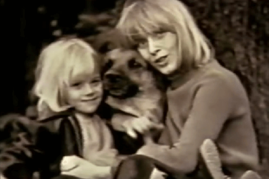 A black-and-white photo of a woman and a young child with blonde hair sitting close together, smiling at the camera while hugging a large dog. The moment appears warm and affectionate, likely from a past decade.